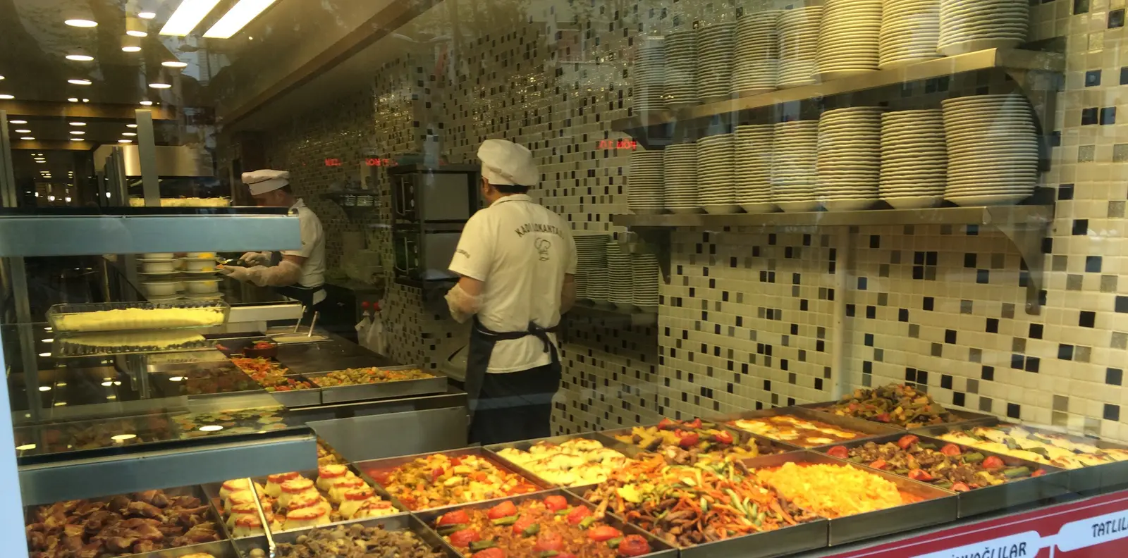 Lokantanın tezgâhı: tepsilerde günün yemekleri, arkada tabak rafları